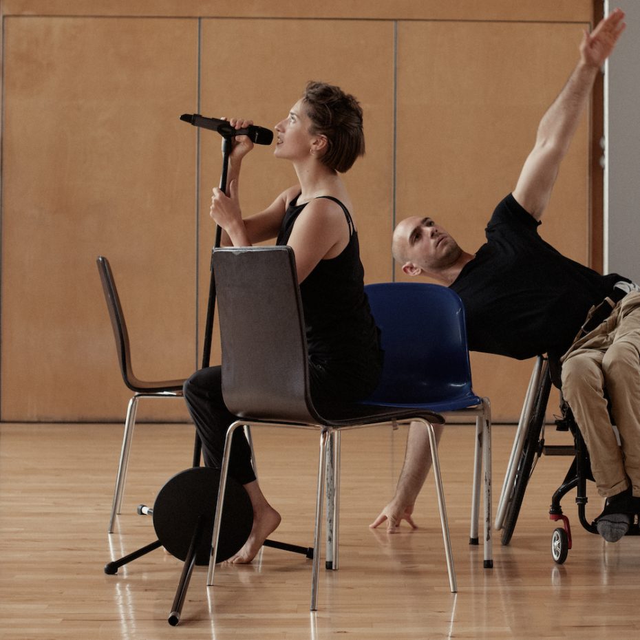Landscape image, 3 chairs, female dancer sits on two of the chairs facing the right side speaking into a microphone. Male dancer behind and to the right is a wheelchair user. He leans to the left making a straight line with his arms from the floor and reaching up to the ceiling. Another female dancer gazes at the scene from the back left corner.
