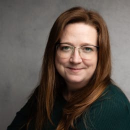 A smiling headshot of a woman with long reddish brown hair and glasses.