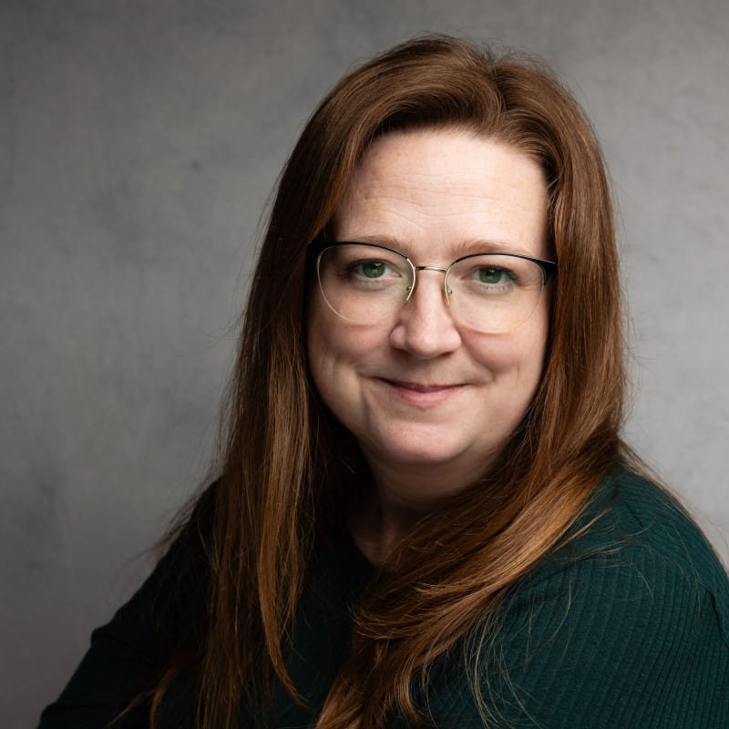 A smiling headshot of a woman with long reddish brown hair and glasses.