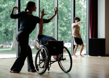 Two dancers, one standing, one using a wheelchair, dance in contact. In the background trees can be seen through the studio windows