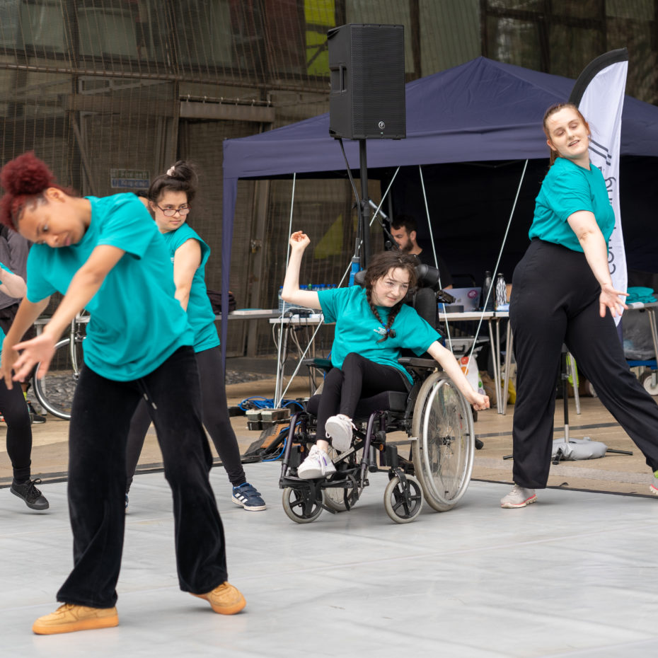Participants dancing outdoors at annual Day of Dance Festival 2022. They wear teal t-shirts and black trousers. They are standing on a dance mat and appear to be are moving towards different directions.