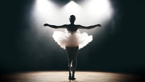 Ballet dancer wearing a tutu. They are standing on stage with arms wide open. The photo is captured from the back.