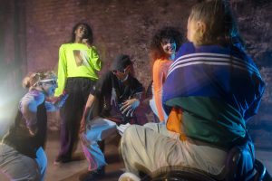 A group of people wearing colourful, vibrant clothing are smiling joyfully as they dance together in a circle. The scene is set against a textured brick wall, adding a rustic backdrop to their cheerful movements and expressive energy. The photograph is taken from a slightly low angle, with the person in the foreground turned away from the camera - adding depth and perspective to the scene.