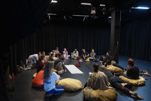 A large group of people, sitting in a circle in a dimly lit indoor space. Some are sitting on bin bags whilst others are lying down. White paper sheets are spread on the floor, along with a box of pens for use during the workshop.