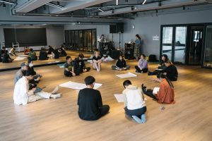 A group of participants engaging in a workshop, all sat on wooden floors with large paper sheets scattered in the middle.