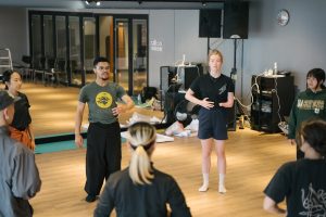 A group of seven participants stand in a circle whilst engaging in a dance workshop, each with one arm raised in front of their abdomen. In the foreground, three individuals face away from the camera, while four others in the background face toward it. The group is positioned near a large mirror in a bright studio space with wooden floors.