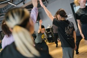 Close-up shot of a dance workshop. The focus is on a person at the back, who is leaning forward with a joyful smile and their arm extended upwards.