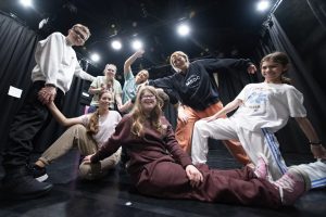 A group of young people smiling and posing backstage. Some are seated on the floor in the foreground, while others stand at the back with their arms raised in celebration. The photo is taken from a low angle, adding a dynamic perspective.