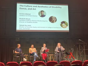 Four people are sat on stage, engaging in a panel talk. Behind them, a large slide is projects onto the wall describing the topic of discuss: ' The Culture and Aesthetics of Disability, Dance and Art' and the names of the panelists.