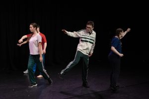 Four young people dancing on stage against a black background. they all face different directions, some balancing on one leg with one arm raised on one side, adding movement and perspective to the scene.