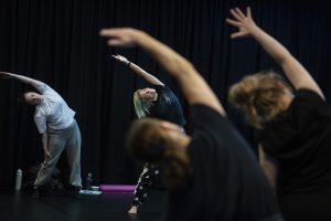 A group of people dancing in a dimly lit indoor space, all leaning to one side with one arm extended overhead. In the foreground, two blurred figures with their backs to the camera enhance the focus on a central dancer in sharp detail.