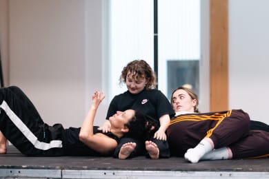 Three people positioned closely together; the central figure is seated and watches the dancer on the left who rests their head on the seated person's lap. The third dancer lies on the right side, looking away. The scene conveys a sense of connection and a feeling of rest.