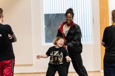 Two dancers smiling joyfully whilst standing in front of one another whilst two people on either side of the frame observe with their arms crossed.