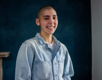 A person wearing a light jeans jacket smiles against a deep blue background. Ania self-describes as follows: "I am a light skinned Latin American, non-binary person. I have a shaved head, big brown eyes and thick dark eyebrows."