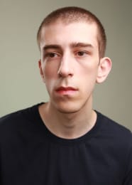 Headshot of a person with short hair, wearing a black shirt, looking away from the camera with a serious expression.