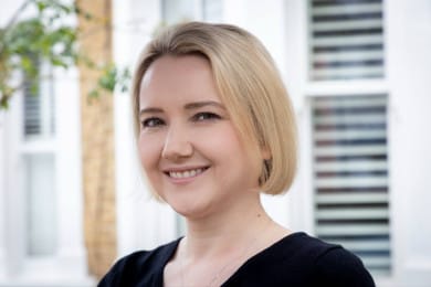 A warm, natural headshot of a person with straight blonde hair styled in a sleek bob cut, smiling gently at the camera. The photo is taken outdoors in soft, natural light, with a blurred background.