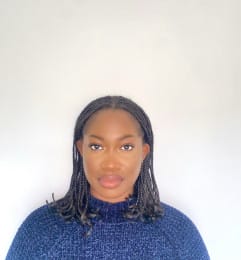 Headshot of a person with short braided hair wearing a blue jumper, looking directly at the camera with a serious expression.