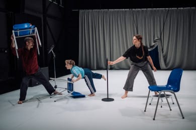 Three performers on a white stage with a grey curtain interact with blue chairs and a microphone stand. One lifts a chair overhead, another balances forward with one leg raised, and a third reaches toward the centre, creating a sense of dynamic movement and tension.