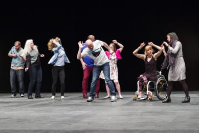 A line of dancers performs on a dark stage, each mid-movement with arms raised or stepping in rhythm. The group includes both standing dancers and a wheelchair user, creating a lively sense of individuality and shared energy.