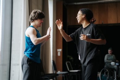 Two people face each other in a dance studio, palms nearly touching in a mirroring exercise, both smiling