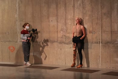 A dancer with a carved wooden leg leans against a concrete wall. A film maker has a camera pointed at the dancer