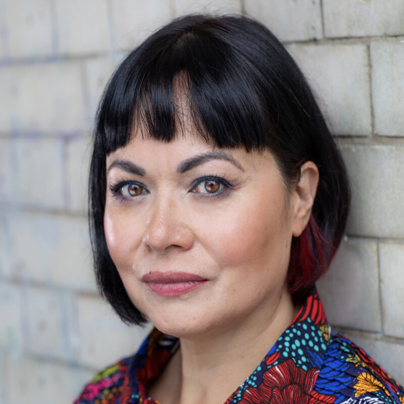 A person with a black bob hairstyle and colourful patterned shirt looks directly at the camera while standing against a light brick wall.