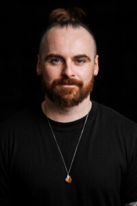 Portrait of a man with a red beard and hair tied in a bun, wearing a black T-shirt, hoop earrings and a pendant necklace, against a black background.