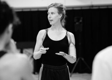 Black-and-white photograph of dancer Annie Hanauer in rehearsal, gesturing with her hands. She wears a black vest top and loose black trousers, with her prosthetic arm visible. Two other dancers are blurred in the foreground.