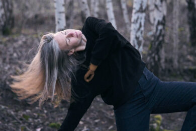 Photograph of dancer Annie Hanauer performing outdoors in a wooded setting. She leans back with her eyes closed and her long hair flowing, with her prosthetic arm visible as she extends one leg behind her.