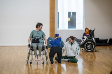 A photograph of three people in a bright dance studio, moving together on a wooden floor. One person is seated in a wheelchair while the others move alongside and support them. Another person is seated in the background beside a large powered wheelchair.
