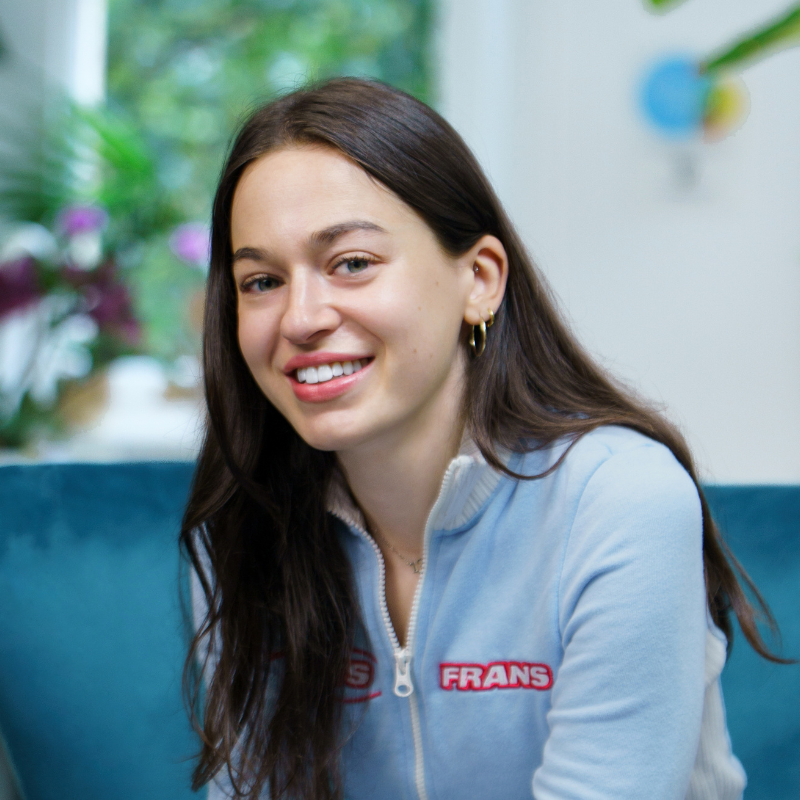 A portrait of a smiling young woman with long dark hair, wearing a light blue zip-up top with red lettering. She is seated indoors on a blue sofa, with plants and a softly blurred, bright background behind her.