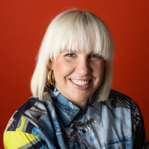 Portrait of Sarah Blanc smiling at the camera against a red background. She has blonde hair with a straight fringe and wears large hoop earrings, a patterned shirt and denim jacket with a chain necklace.