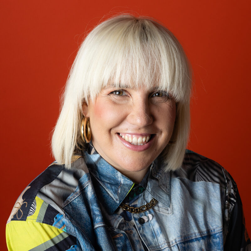 Portrait of Sarah Blanc smiling at the camera against a red background. She has blonde hair with a straight fringe and wears large hoop earrings, a patterned shirt and denim jacket with a chain necklace.