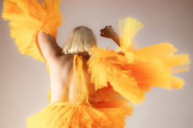 A person seen from behind wearing a voluminous bright yellow-orange tulle costume, with their arms raised and layers of fabric billowing out around them against a neutral background.