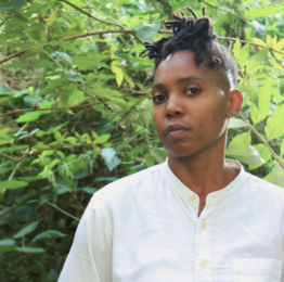 A portrait of a person with short, textured locs, wearing a white buttoned shirt and standing outdoors surrounded by lush green foliage. They look towards the camera with a calm, thoughtful expression.