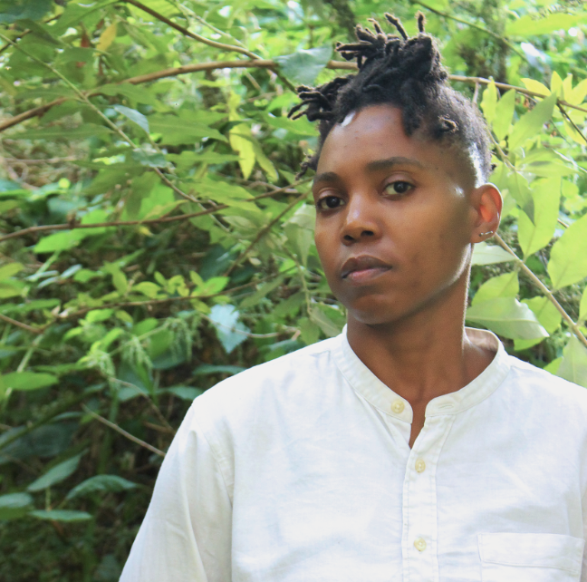 A portrait of a person with short, textured locs, wearing a white buttoned shirt and standing outdoors surrounded by lush green foliage. They look towards the camera with a calm, thoughtful expression.